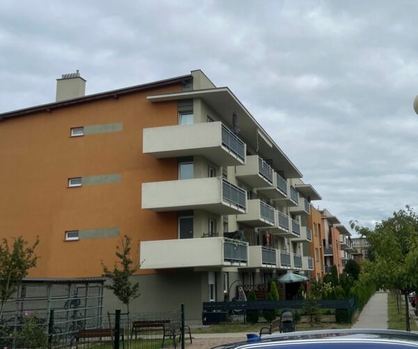 Nowoczesny, pomarańczowy budynek wielorodzinny z balkonami, widziany z perspektywy ulicy. Widoczne drzewa i chodnik. Pochmurne niebo w tle.