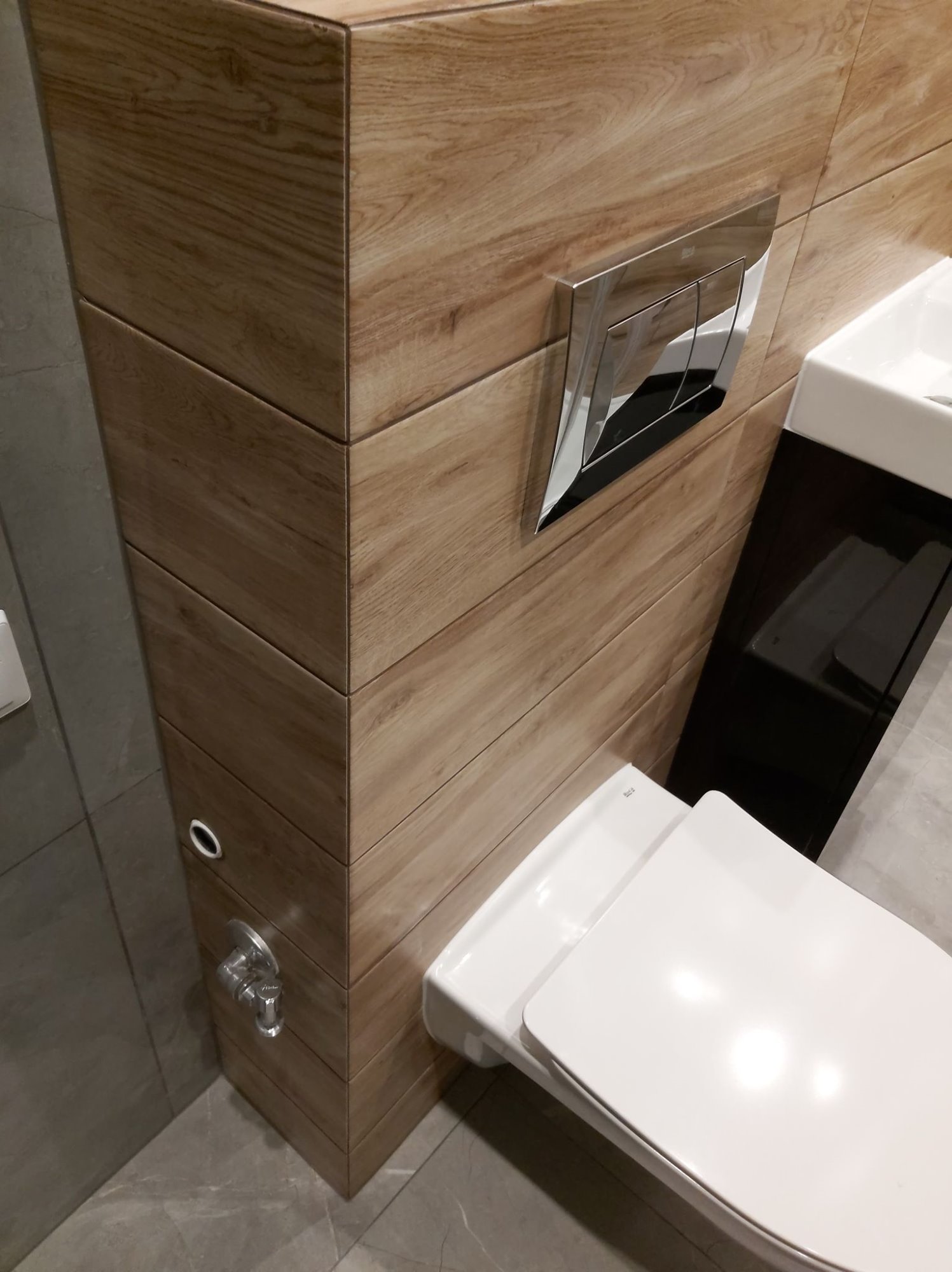 Nowoczesna toaleta podwieszana z drewnopodobną obudową i chromowanym przyciskiem spłukującym, widoczna umywalka z czarną szafką, szare płytki podłogowe.