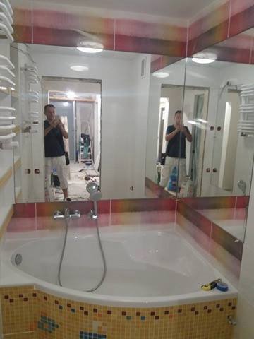 Łazienka w trakcie remontu: widoczna wanna z mozaiką, baterią i słuchawką prysznicową; w lustrze odbija się mężczyzna robiący zdjęcie i fragment pomieszczenia z drabiną.