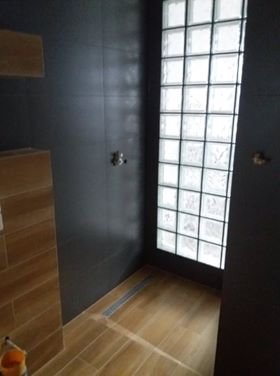 Ciemnoszara łazienka z kabiną prysznicową oddzieloną ścianką z luksferów, podłoga wyłożona płytkami imitującymi drewno, widoczny odpływ liniowy.
