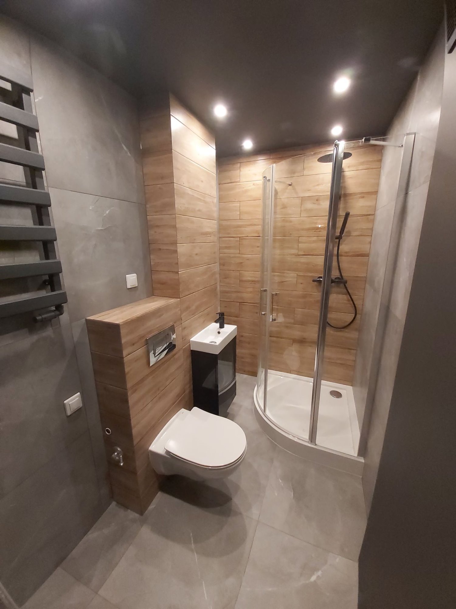 Nowoczesna łazienka z kabiną prysznicową narożną, umywalką nablatową na czarnej szafce, podwieszaną toaletą z zabudową z drewnopodobnych paneli i szarymi płytkami na ścianach i podłodze.