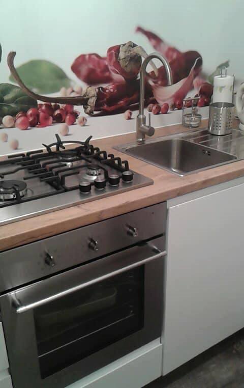 Nowoczesna kuchnia z zabudową: srebrny piekarnik, płyta gazowa i zlewozmywak ze stali nierdzewnej, drewniany blat i białe szafki, ściana ozdobiona fototapetą z motywem papryczek chili.