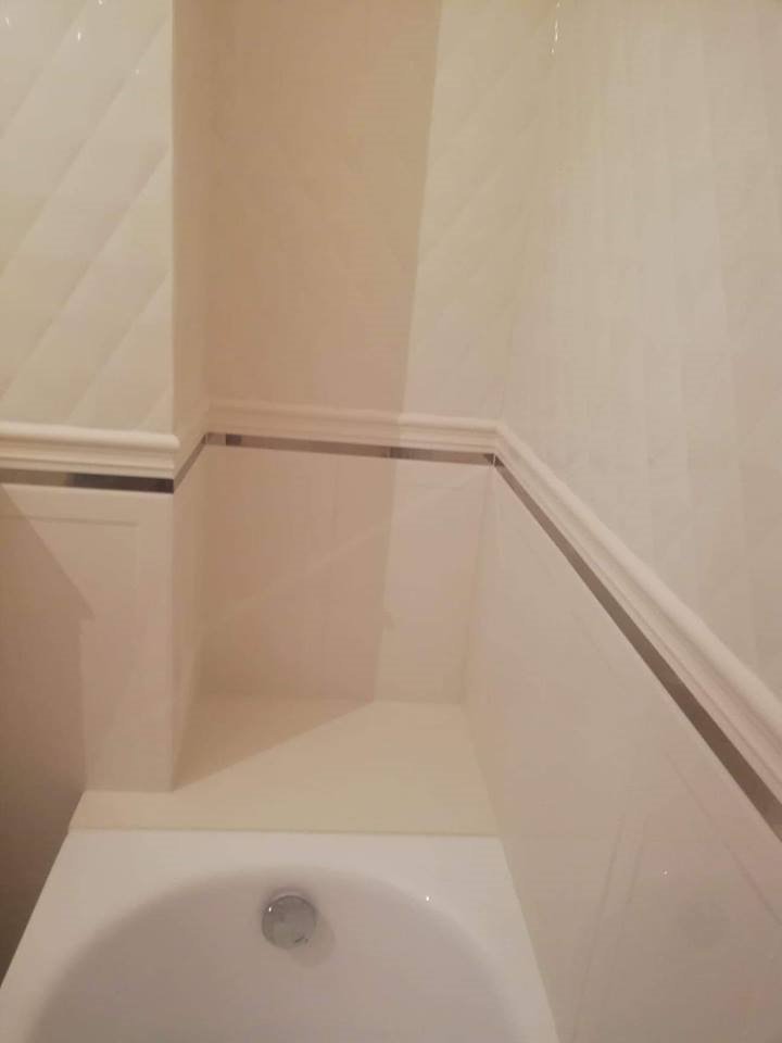 Wykończona łazienka z białymi płytkami, dekoracyjnym gzymsem z metalową wstawką i widoczną wanną.