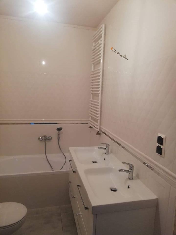 Jasna łazienka z wanną, umywalką z dwiema misami i grzejnikiem drabinkowym, wykończona panelami ściennymi z delikatnym wzorem rombów.