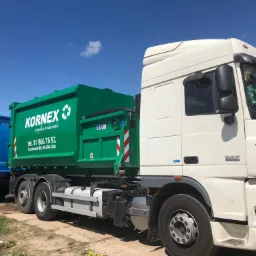 Ciężarówka z zielonym kontenerem firmy KORNEX z numerem telefonu i logiem, zaparkowana na placu, obok niebieski kontener na przyczepie, błękitne niebo w tle.