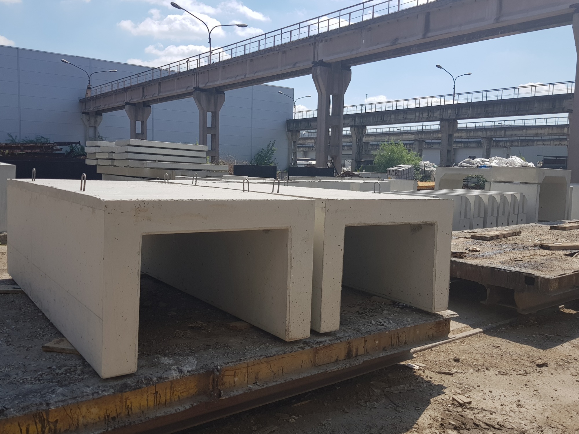 Prefabrykowane betonowe elementy tunelowe typu U na placu, widoczne metalowe uchwyty montażowe, w tle konstrukcja mostowa i hala przemysłowa.