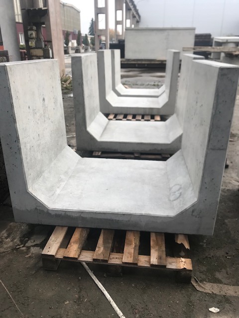 Betonowe korytka ściekowe U-kształtne, ułożone na drewnianych paletach, widoczne na zewnątrz, w tle budynki przemysłowe.