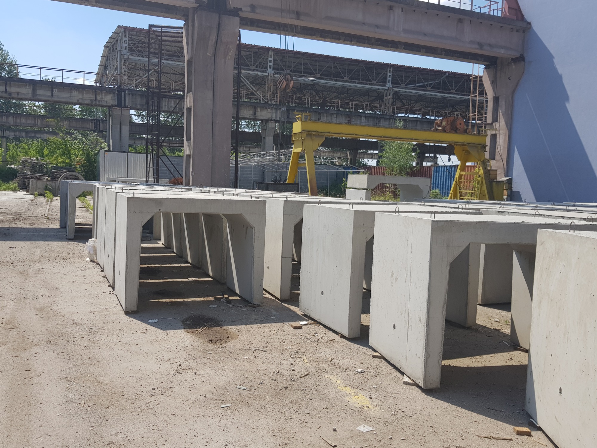 Betonowe prefabrykaty w kształcie litery U, ustawione rzędem na placu budowy z dźwigiem bramowym w tle.