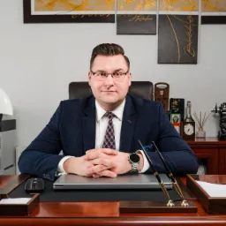 Mężczyzna w garniturze siedzi przy biurku z laptopem, w tle maszyna do pisania i dekoracyjne obrazy.