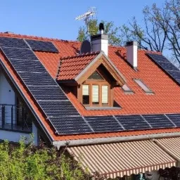 Dom jednorodzinny z czerwonym dachem pokrytym panelami słonecznymi, widoczne okno dachowe i komin, antena telewizyjna na dachu.