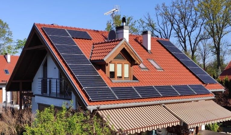 Dom jednorodzinny z czerwonym dachem pokrytym panelami słonecznymi, widoczne okno dachowe i komin, antena telewizyjna na dachu.