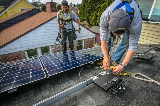 Dwóch monterów w uprzężach zabezpieczających instaluje panele na dachu domu jednorodzinnego, jeden z nich łączy przewody do inwertera.