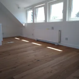 Jasne, przestronne poddasze z nowo ułożonym parkietem, białymi ścianami i grzejnikiem pod trzema oknami dachowymi, z promieniami słońca padającymi na podłogę.