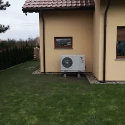 Biała pompa ciepła Rotenso zamontowana na betonowych bloczkach przy ścianie domu z brązową dachówką, widok z boku na trawnik i żywopłot.