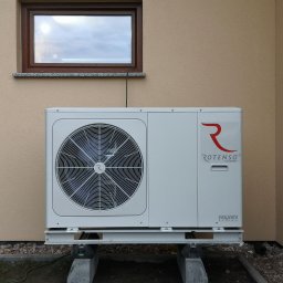 HydroRaaD - Biała pompa ciepła Rotenso Aquami Series, zamontowana na betonowych podstawach przy ścianie budynku z oknem. Widoczny wentylator i logo firmy.