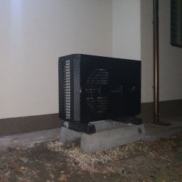 HydroRaaD - Czarna pompa ciepła typu monoblok zainstalowana na betonowym fundamencie przy elewacji budynku, widok z niskiej perspektywy, jesienne otoczenie z liśćmi.