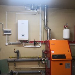 HydroRaaD - Pomieszczenie techniczne z zainstalowanym kotłem gazowym UltraGas, białym zasobnikiem ciśnieniowym, oraz licznymi rurami instalacji grzewczej i wodnej. Na ścianie widoczny biały piec Kaisai.