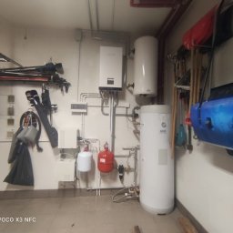 HydroRaaD - Białe zbiorniki i piec gazowy Netsu zamontowane w garażu, widoczne rury, zabezpieczenia i narzędzia ogrodowe na ścianach.