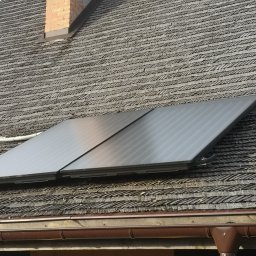 HydroRaaD - Dwa panele słoneczne na dachu krytym gontem drewnianym, widoczne rynny i komin z cegły.