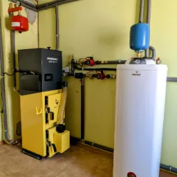 Żółto-czarny piec na paliwo stałe marki Pereko Q-Pell EVO 100, obok biały bojler Galmet z niebieskim zbiornikiem wyrównawczym, orurowanie grzewcze w izolacji na żółtej ścianie, brązowe płytki...