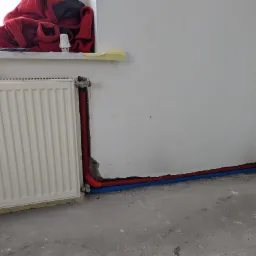 Biały grzejnik panelowy z podłączonymi rurami grzewczymi w kolorze czerwonym i niebieskim, prowadzącymi wzdłuż ściany w trakcie montażu, widoczne niedoskonałości w wykończeniu ściany i podłogi.