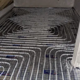 Rozłożone białe rury ogrzewania podłogowego na folii z siatką, widoczne mocowania rur i etykiety 'MONTER quality', surowy stan poddasza z oknem i fragmentem drewnianej konstrukcji dachu.