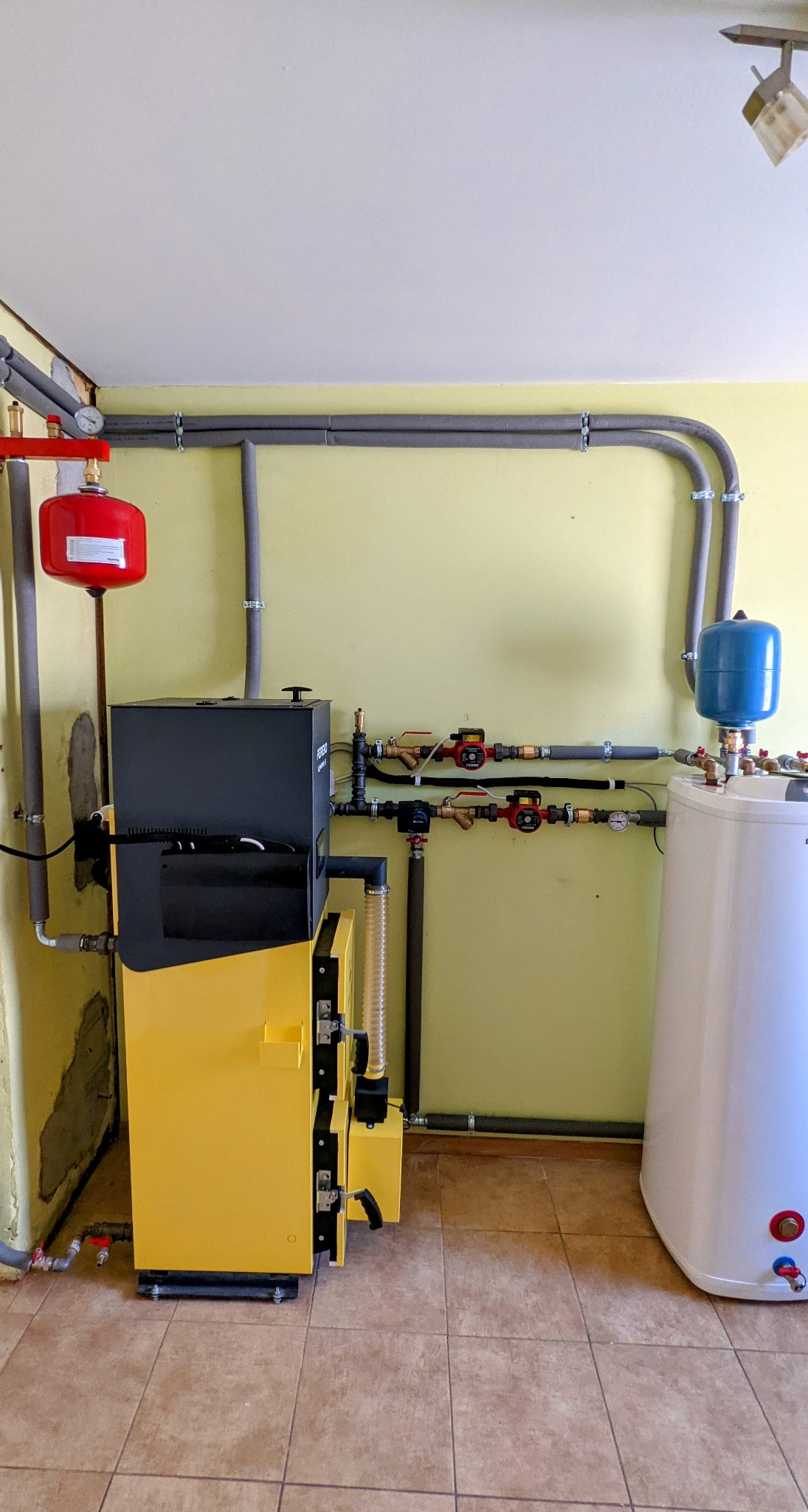 Żółto-czarny piec na paliwo stałe z podłączonymi instalacjami: rurami, zbiornikami wyrównawczymi (czerwonym i niebieskim) oraz białym zasobnikiem ciepłej wody użytkowej. Widoczne liczne zawory...
