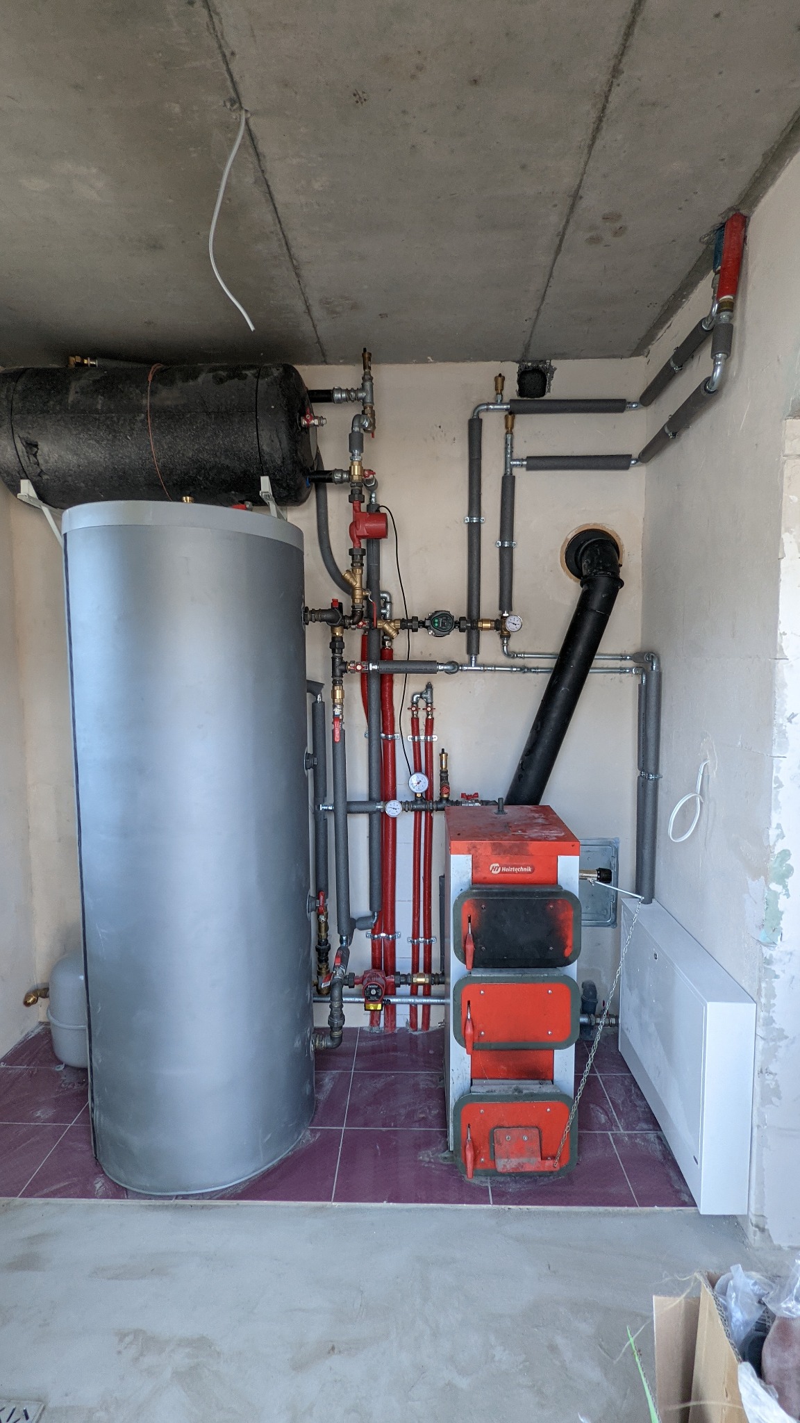 Instalacja kotła na paliwo stałe Heiztechnik z zasobnikiem ciepłej wody użytkowej, widoczne rury grzewcze z izolacją, manometry i zawory, pomieszczenie w trakcie wykańczania.