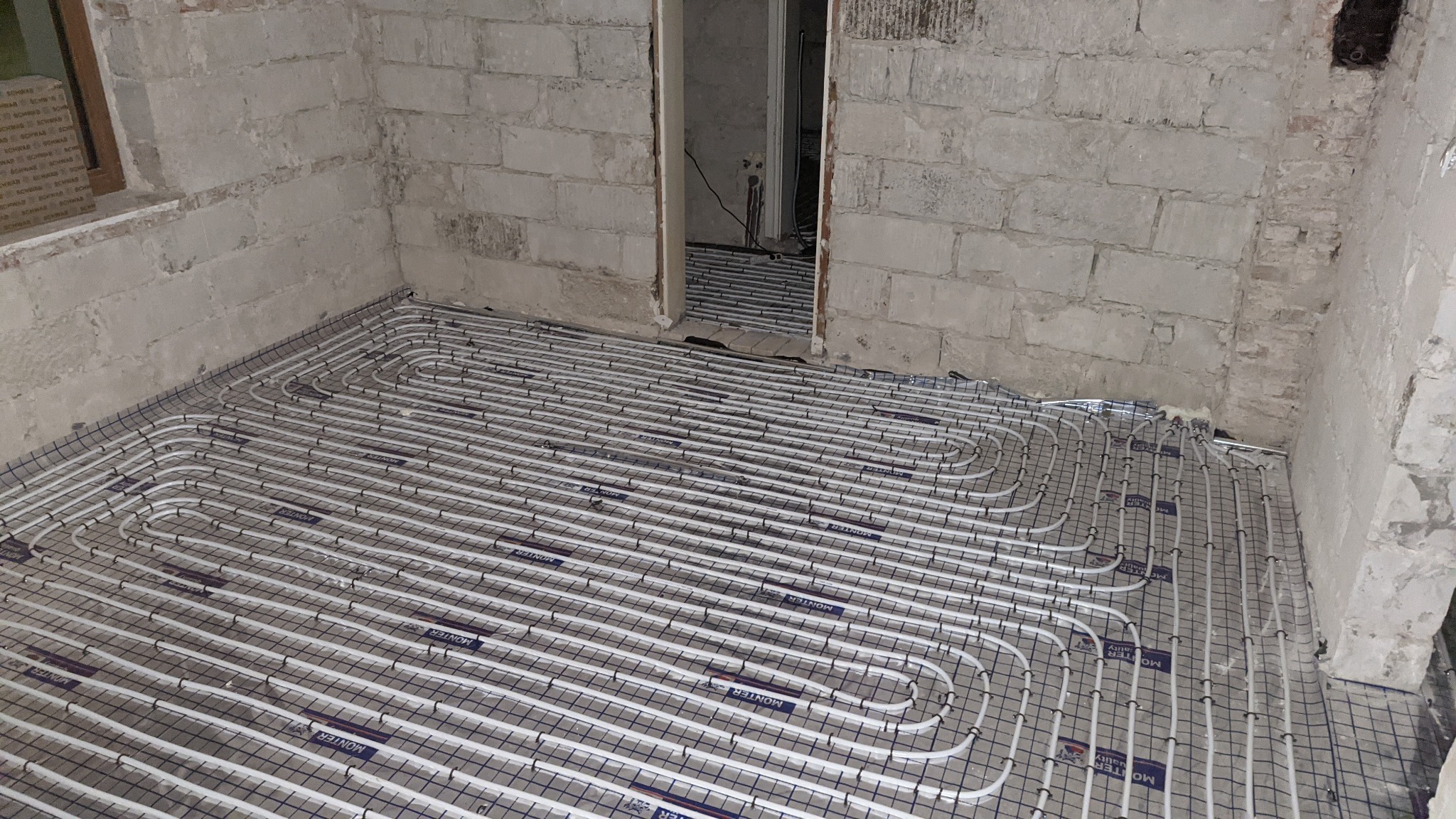 Białe rury ogrzewania podłogowego ułożone na siatce montażowej w surowym pomieszczeniu z ceglanymi ścianami przed wylewką.