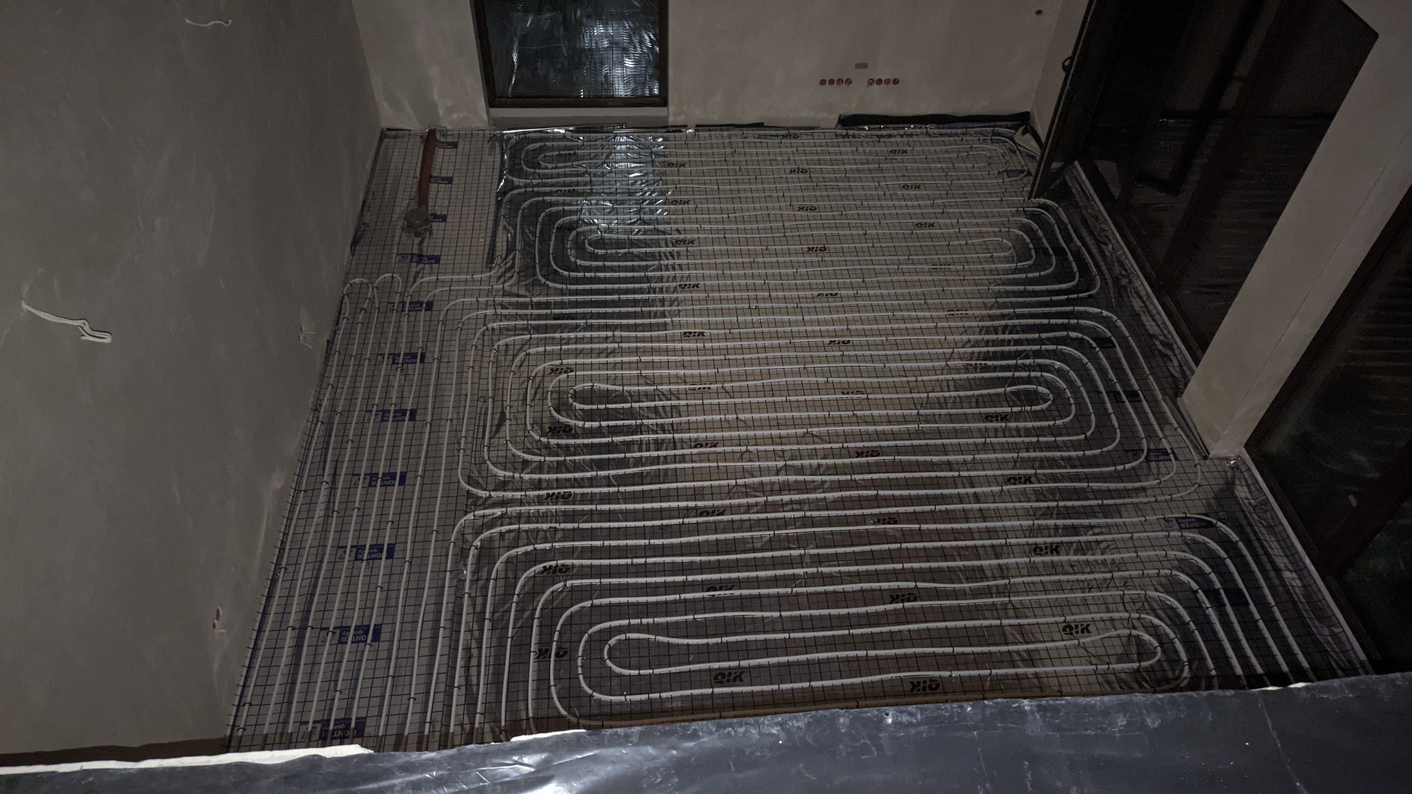 Montaż ogrzewania podłogowego: białe rurki rozprowadzone na czarnej siatce, widok z góry na instalację przed zalaniem wylewką.