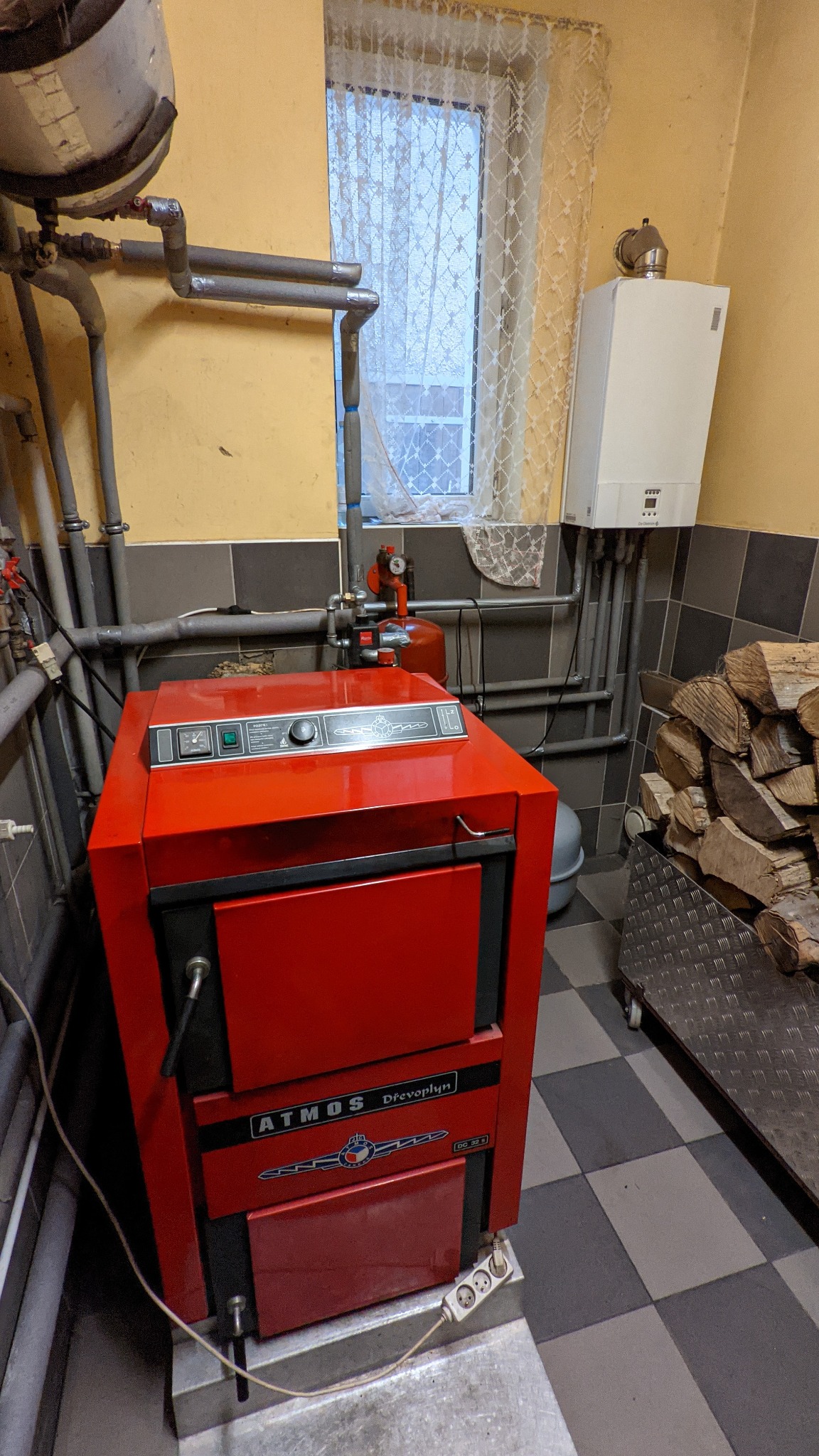 Czerwony piec na paliwo stałe Atmos Drevo Plyn DG 32 S, widoczny w kotłowni w Kolimagach, z instalacją grzewczą i zapasem drewna na wózku.