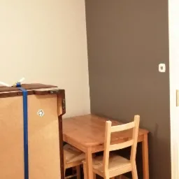 Ściana pomalowana na dwa kolory: beżowy i szary, drewniany stół z krzesłami, widoczny fragment mebla zabezpieczonego niebieską taśmą.