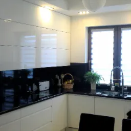 Nowoczesna kuchnia z białymi, lakierowanymi szafkami, czarnym blatem i backsplash, okno z roletami dzień-noc, ozdobne rośliny doniczkowe i koszyk z owocami.