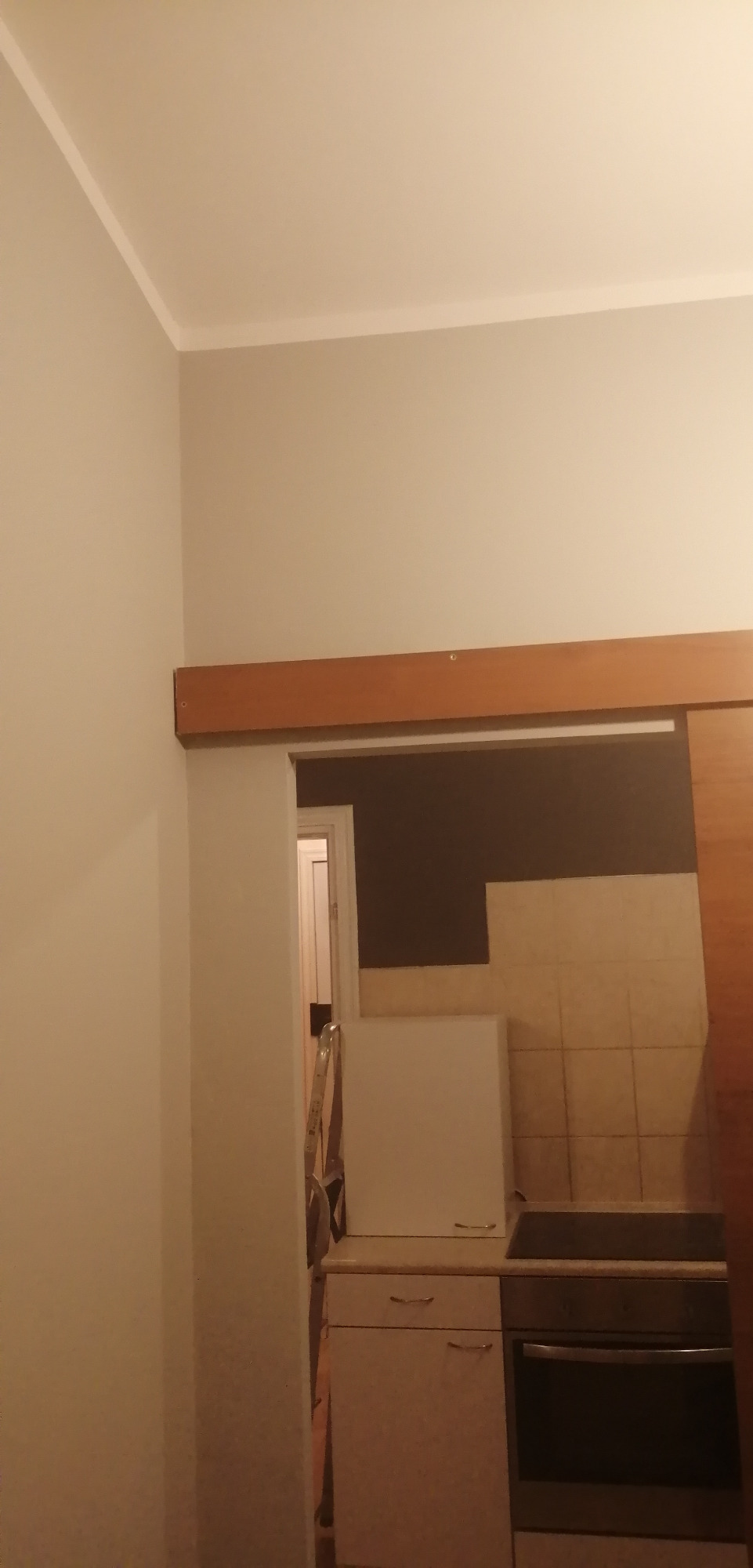 Fragment pokoju z widocznym sufitem, ścianą pomalowaną na jasny kolor oraz drewnianą belką nad drzwiami, w tle widoczna kuchnia z płytkami i sprzętem.