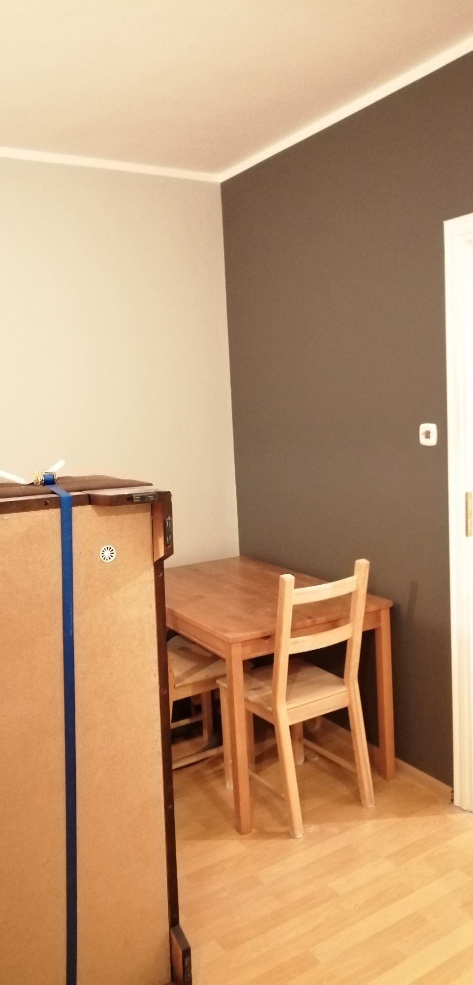 Ściana pomalowana na dwa kolory: beżowy i szary, drewniany stół z krzesłami, widoczny fragment mebla zabezpieczonego niebieską taśmą.