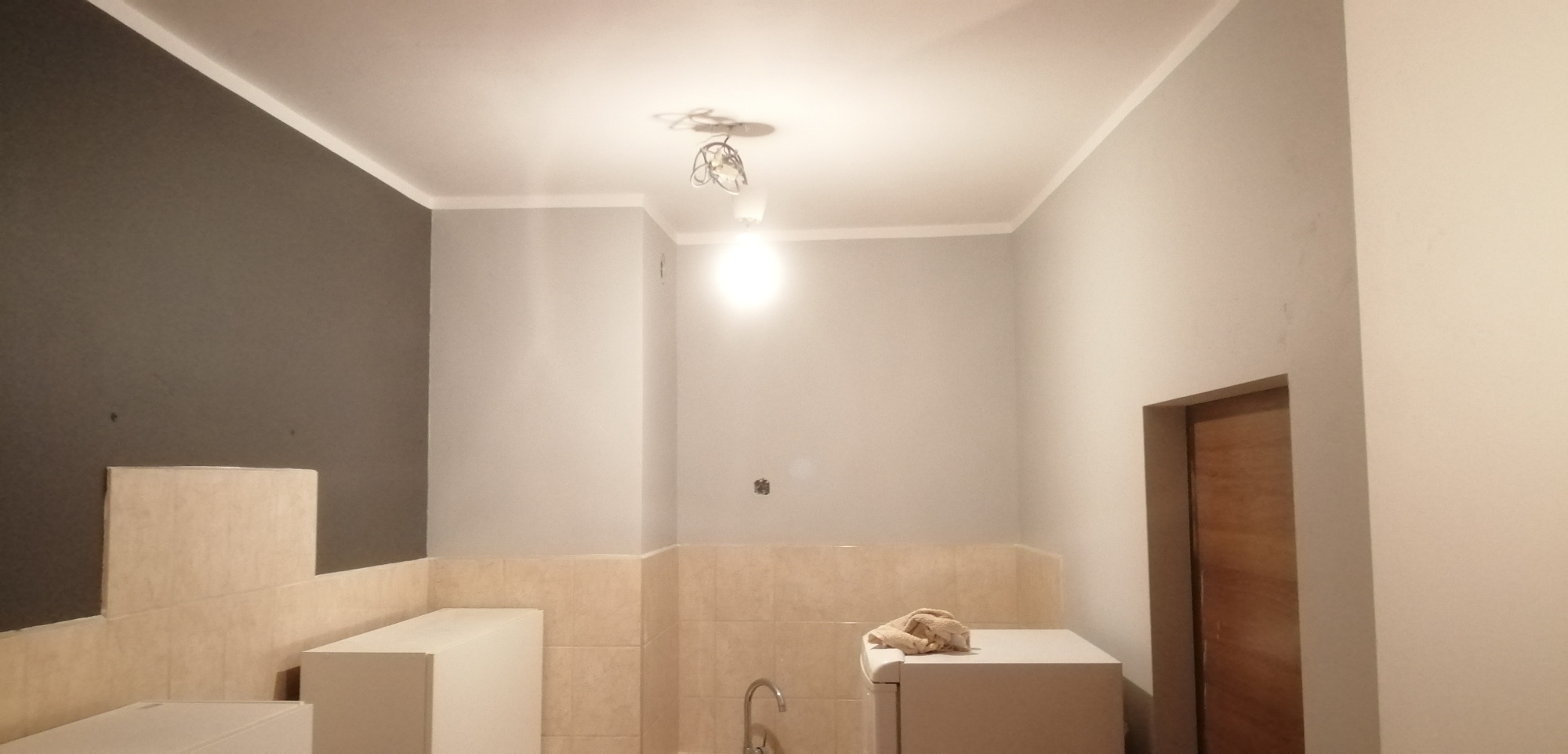 Wnętrze kuchni w trakcie remontu, z szarą ścianą, beżowymi płytkami na ścianach, białymi szafkami i widoczną instalacją oświetleniową na suficie. Widoczne ślady po gniazdkach elektrycznych.