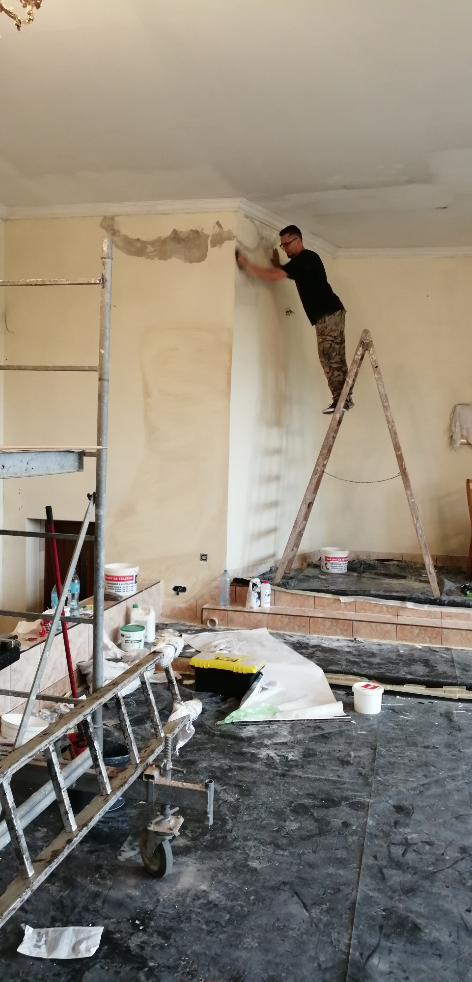 Mężczyzna w okularach, stojący na drewnianej drabinie, wyrównuje szpachlą ścianę z widocznymi ubytkami, w tle rusztowanie, wiaderka z farbą i narzędzia na podłodze zabezpieczonej folią malarską.