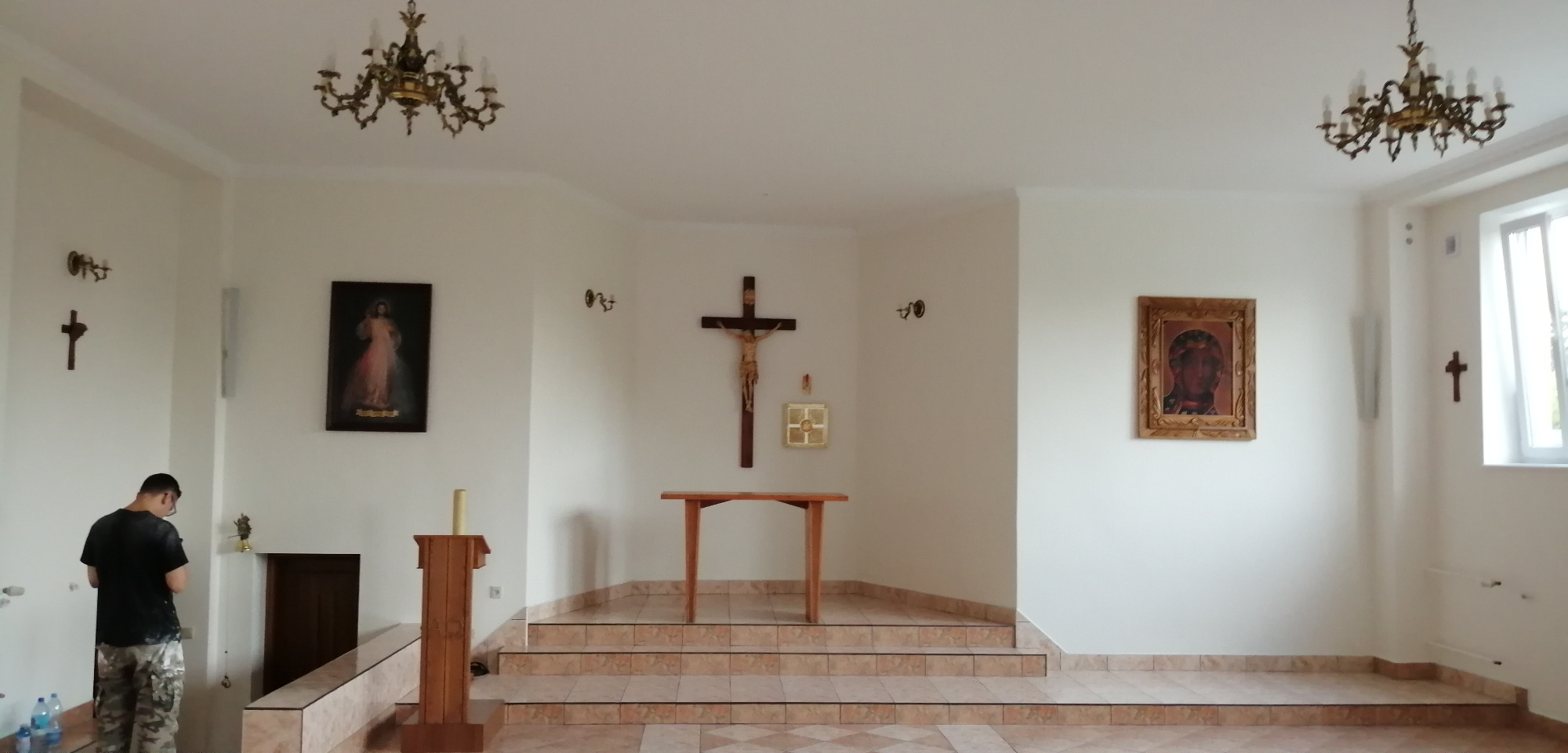 Wnętrze kaplicy po remoncie, z nową, jasną kolorystyką ścian i podłogi wyłożonej płytkami w beżowo-brązową szachownicę, oświetlone przez dwa żyrandole w stylu retro, z obrazami i krucyfiksem...