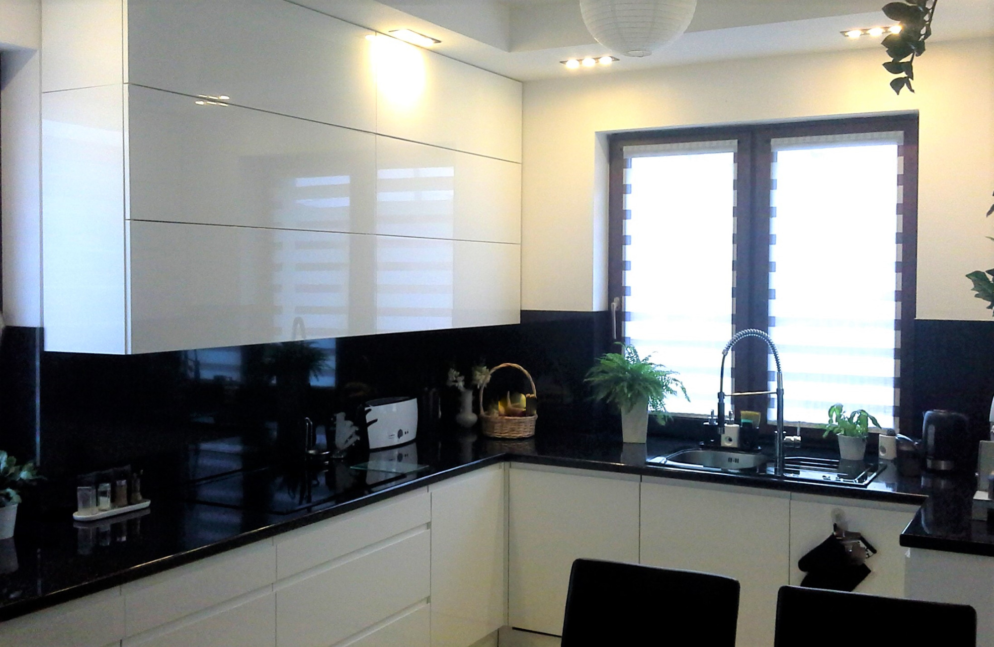 Nowoczesna kuchnia z białymi, lakierowanymi szafkami, czarnym blatem i backsplash, okno z roletami dzień-noc, ozdobne rośliny doniczkowe i koszyk z owocami.