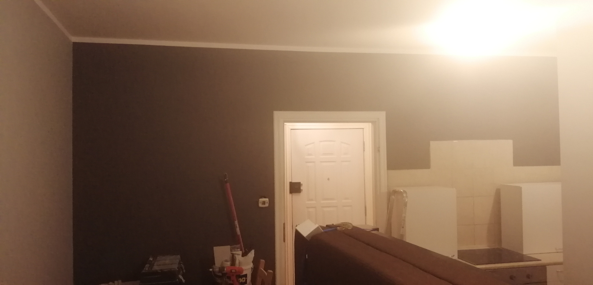 Pokój po malowaniu: ściana w odcieniach brązu kontrastuje z białą listwą sufitową i drzwiami. Widoczne narzędzia malarskie i elementy wyposażenia wnętrza.