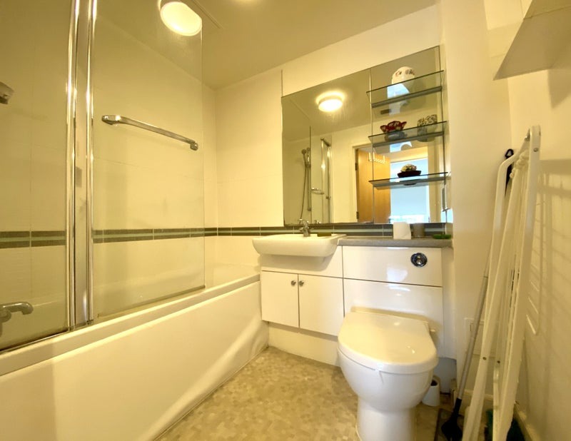 Jasna łazienka z wanną za szklanym parawanem, umywalką z szafką, toaletą i lustrem z półkami, na których stoją dekoracje.