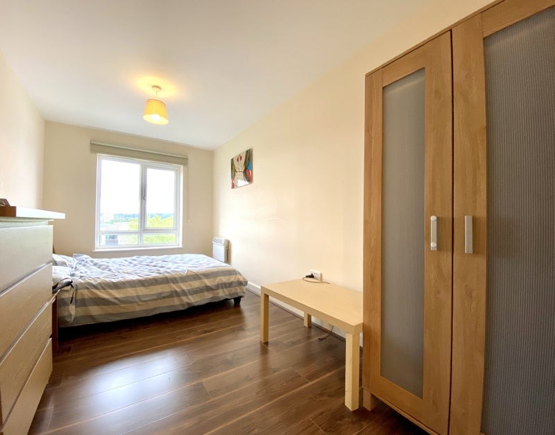 Jasne wnętrze sypialni z drewnianą podłogą, szafą z przeszkleniami, łóżkiem w paski i małym stolikiem, widok na miasto za oknem.