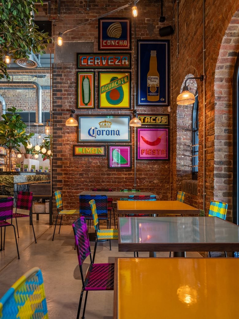 Wnętrze restauracji w stylu meksykańskim z ceglanymi ścianami, kolorowymi krzesłami i plakatami reklamowymi napojów i jedzenia. Oświetlenie w postaci żarówek edisona.