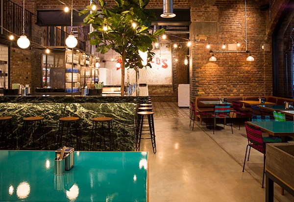 Wnętrze lokalu gastronomicznego z barem z zielonego marmuru, drewnianymi stołkami barowymi, ceglanymi ścianami, rośliną doniczkową i dekoracyjnym oświetleniem.