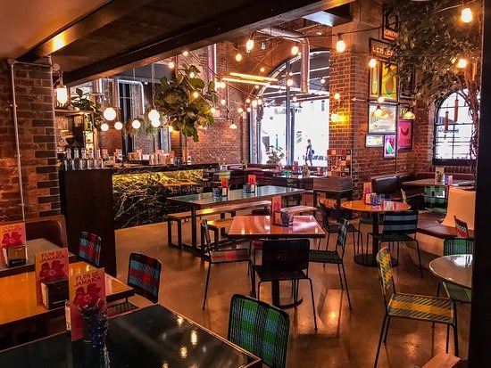 Wnętrze restauracji z ceglanymi ścianami, marmurowym barem i kolorowymi krzesłami, oświetlone wiszącymi lampami i dekoracjami roślinnymi.