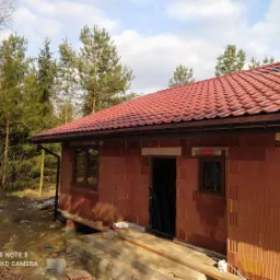 Dom murowany w trakcie budowy, z czerwonym dachem z dach&oacute;wki ceramicznej, widoczne okna z brązowymi ramami i ściany z czerwonej cegły, tło stanowią drzewa iglaste i liściaste.