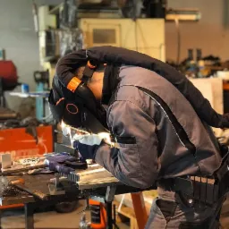 Pracownik w kombinezonie ochronnym spawa metalowe elementy na stole warsztatowym, używając maski spawalniczej z systemem wentylacji i filtrem powietrza.