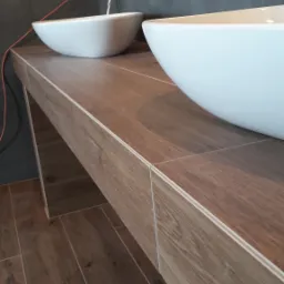 Dwie białe, owalne umywalki nablatowe z chromowaną armaturą na blacie z płytek imitujących drewno. Szare płytki na ścianie i podświetlenie LED. Widoczny fragment pomarańczowego kabla.