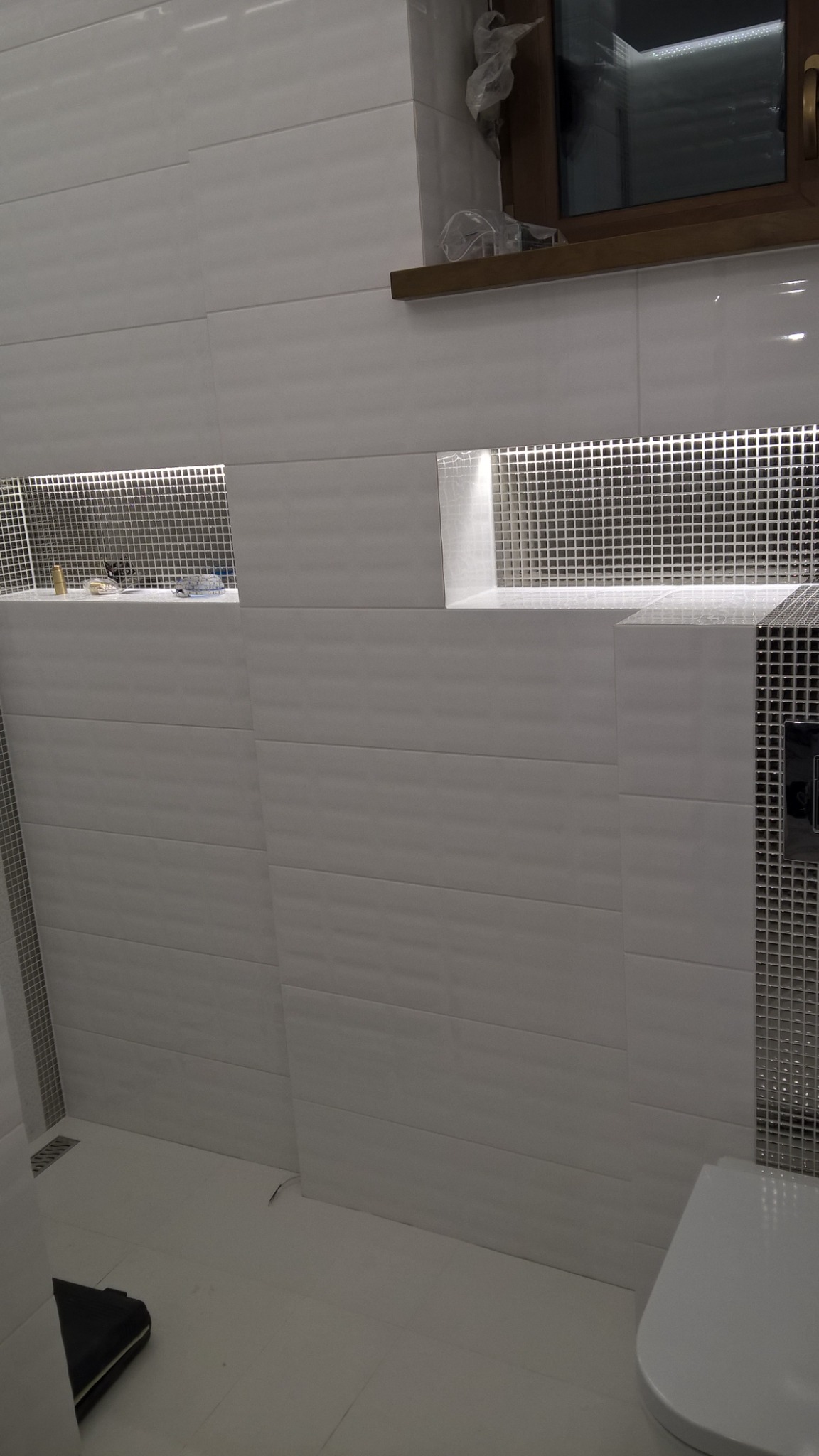 Biała łazienka z poziomymi, teksturowanymi płytkami, wnękami z mozaiką i oświetleniem LED, widoczne elementy toalety i okno z drewnianą ramą.