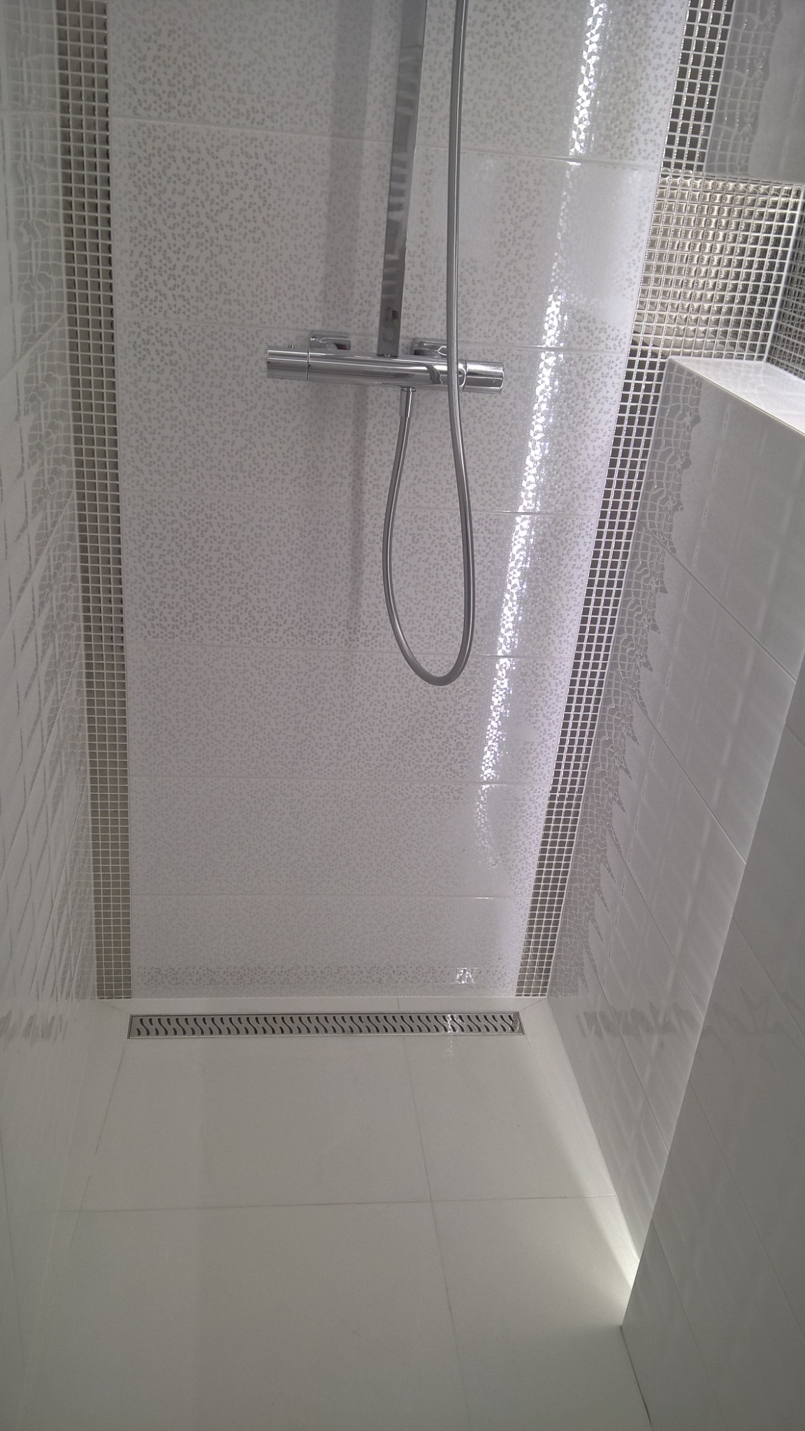 Nowoczesna kabina prysznicowa z białymi, wzorzystymi płytkami i odpływem liniowym, wykończona mozaiką. Widoczna chromowana bateria i deszczownica.